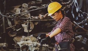 Woman Wears Yellow Hard Hat Holding Vehicle Part